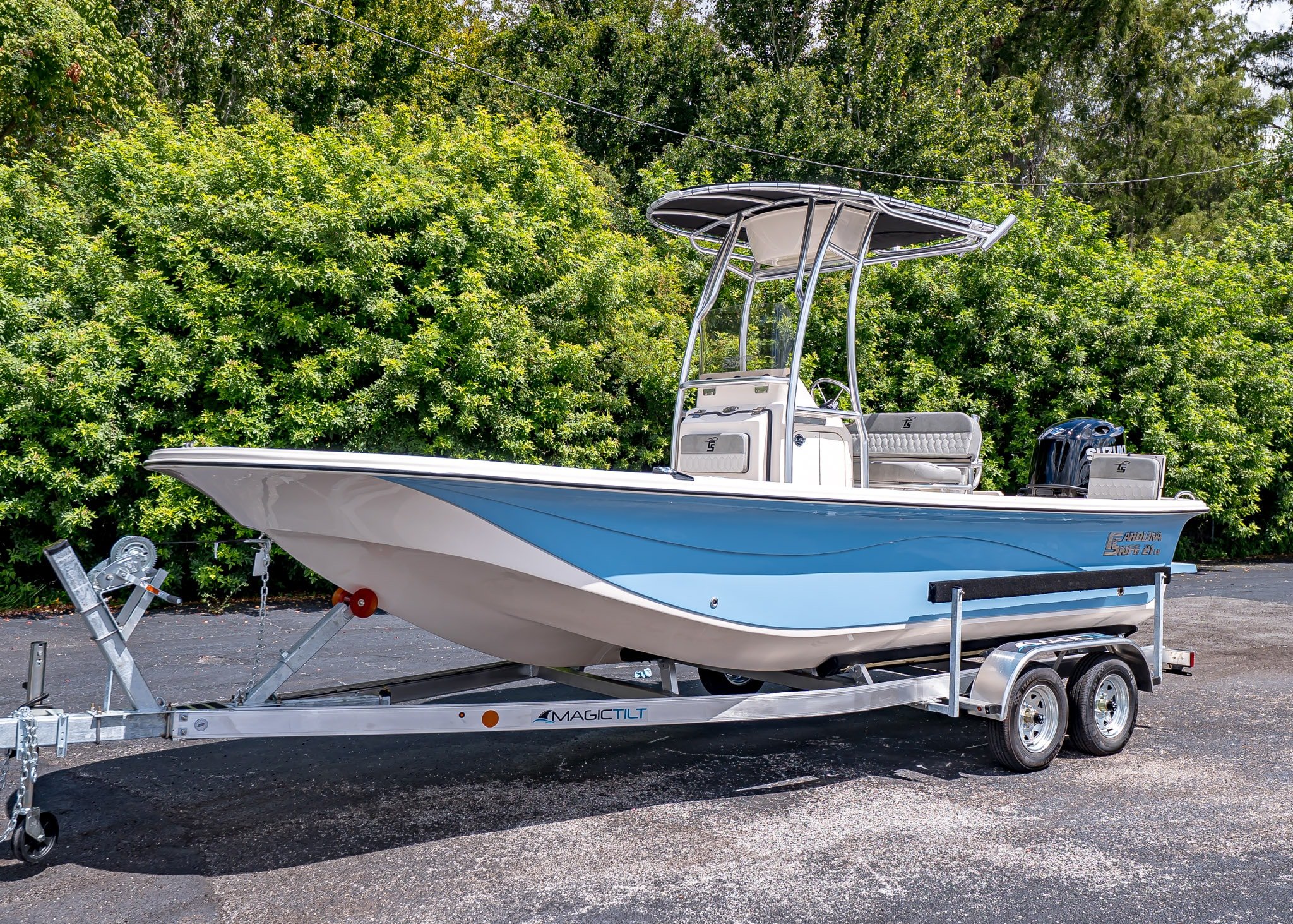 A light blue and white Carolina Skiff, the best entry boat, sits on a dual-axle trailer parked on pavement with green bushes and trees in the background.